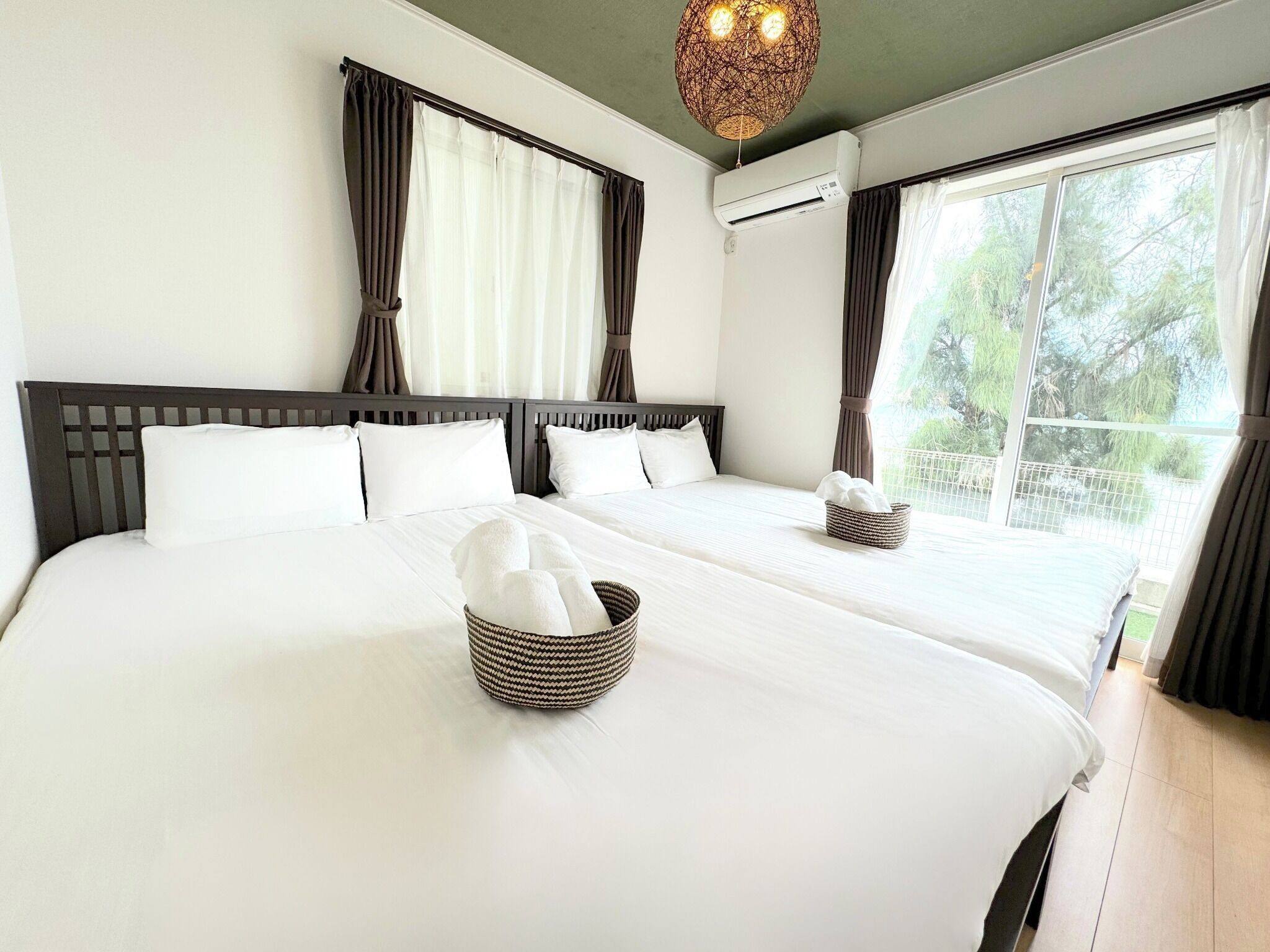 A hotel room with two beds, a large window with curtains, a ceiling light fixture, and an air conditioning unit.