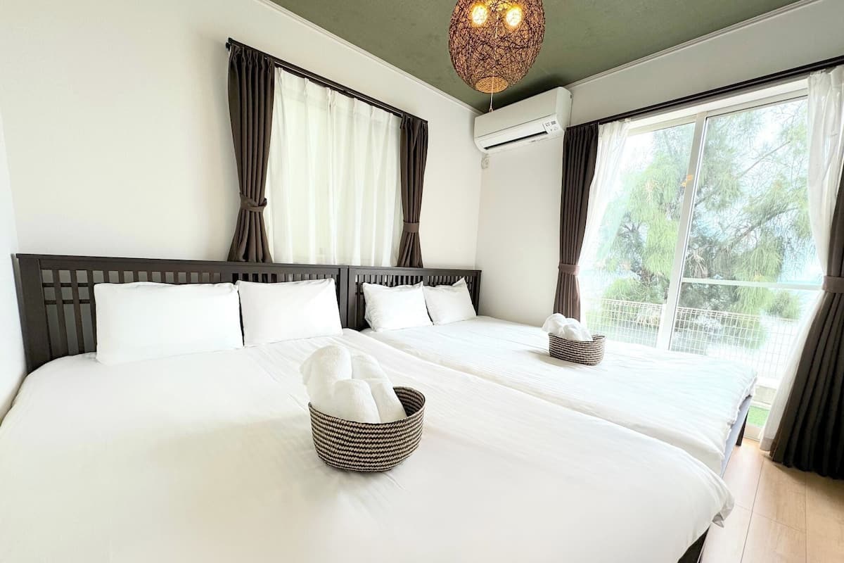 A hotel room with two beds, a large window with curtains, an air conditioning unit, and a woven basket with towels.