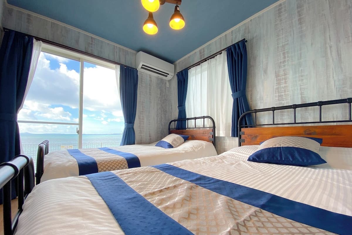 A hotel room with two beds, a large window with curtains, an air conditioning unit, and a view of the sea.
