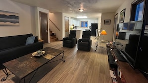 A living room with a sofa, two armchairs, a coffee table, and a TV. There is a kitchen in the background with white cabinets and a black refrigerator. - Mountain Views,Downtown,RMNP,Fenced Backyard,Pet Friendly,Hot Tub!Woods&Waves (Estes Park)