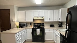 A compact kitchen with white cabinets, a black stove, a microwave, a refrigerator, and a coffee maker. - Mountain Views,Downtown,RMNP,Fenced Backyard,Pet Friendly,Hot Tub!Woods&Waves (Estes Park)