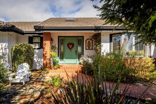 A house with a green door, a brick front porch, and a garden with various plants and a white chair.