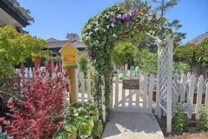 A garden entrance with a white picket fence, a yellow sign, and a variety of plants and flowers.