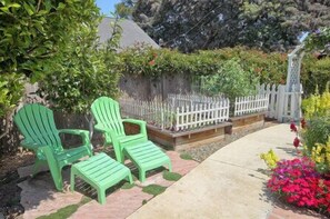 Relax in a serene outdoor space surrounded by lush plants and vibrant flowers, with a comfortable seating area and a wooden deck in the background.