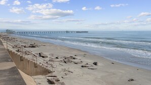 A beach with a concrete staircase leading down to the shore, a pier extending into the water, and a clear sky with scattered clouds. - New Heated Pool & HOT tub! Close Ocean Views! (Galveston)