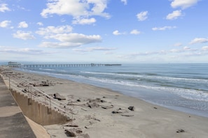 Una playa con una escalera de hormigón que baja hasta la orilla, un muelle que se adentra en el agua, y un cielo despejado con algunas nubes.