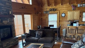 A cozy cabin interior with a stone fireplace, wooden walls, a dining area, and a living room furnished with leather couches and a coffee table.