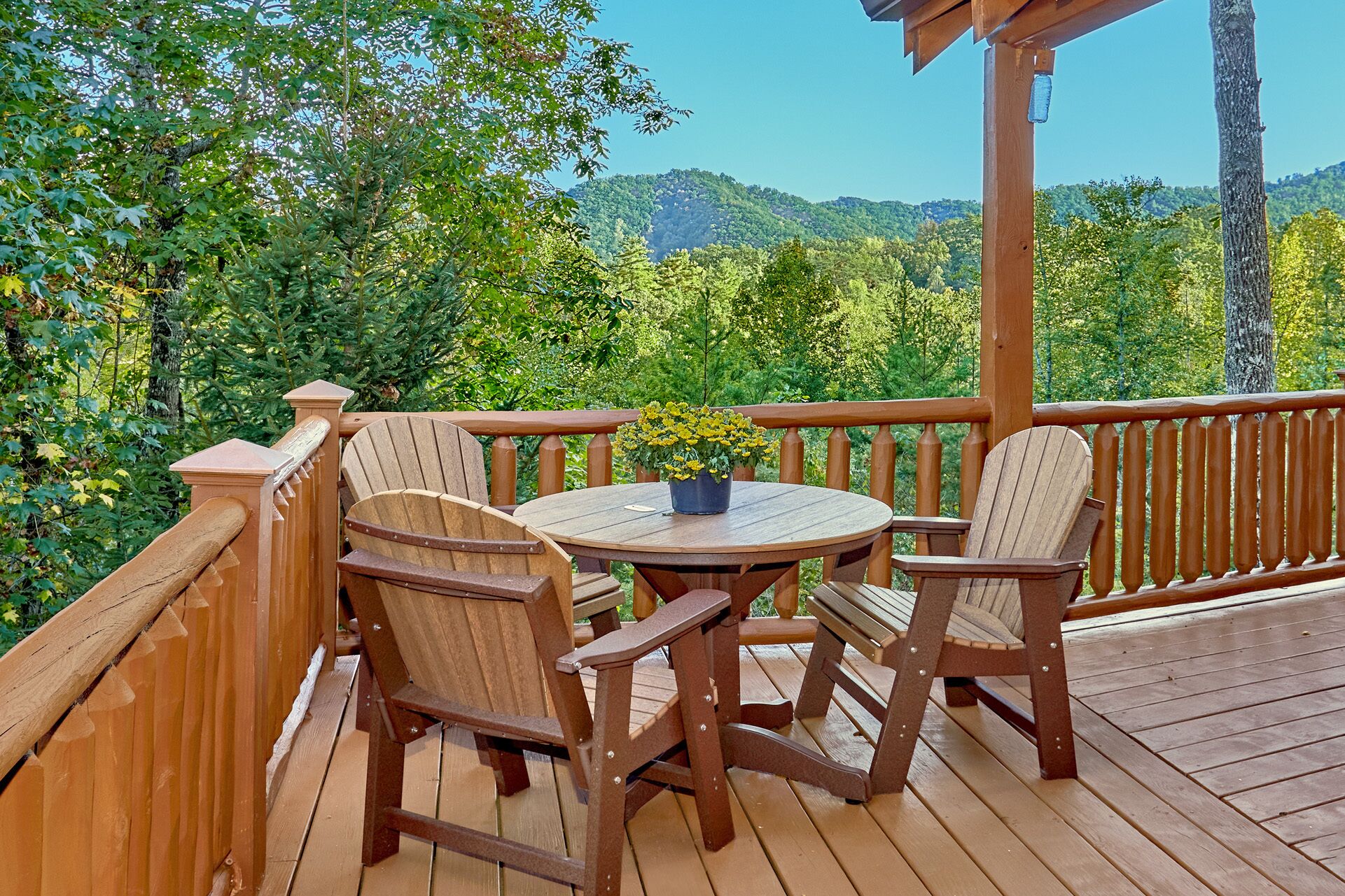 Una terraza de madera con una mesa redonda y cuatro sillas Adirondack, rodeada por una barandilla de madera.