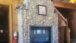 A stone fireplace with a framed sign above it, wooden beams on the ceiling, and a wooden staircase to the right. - Cozy Mountain Retreat / Great For Reunions, Family Retreats,  Room For Tents! (Duck Creek Village)