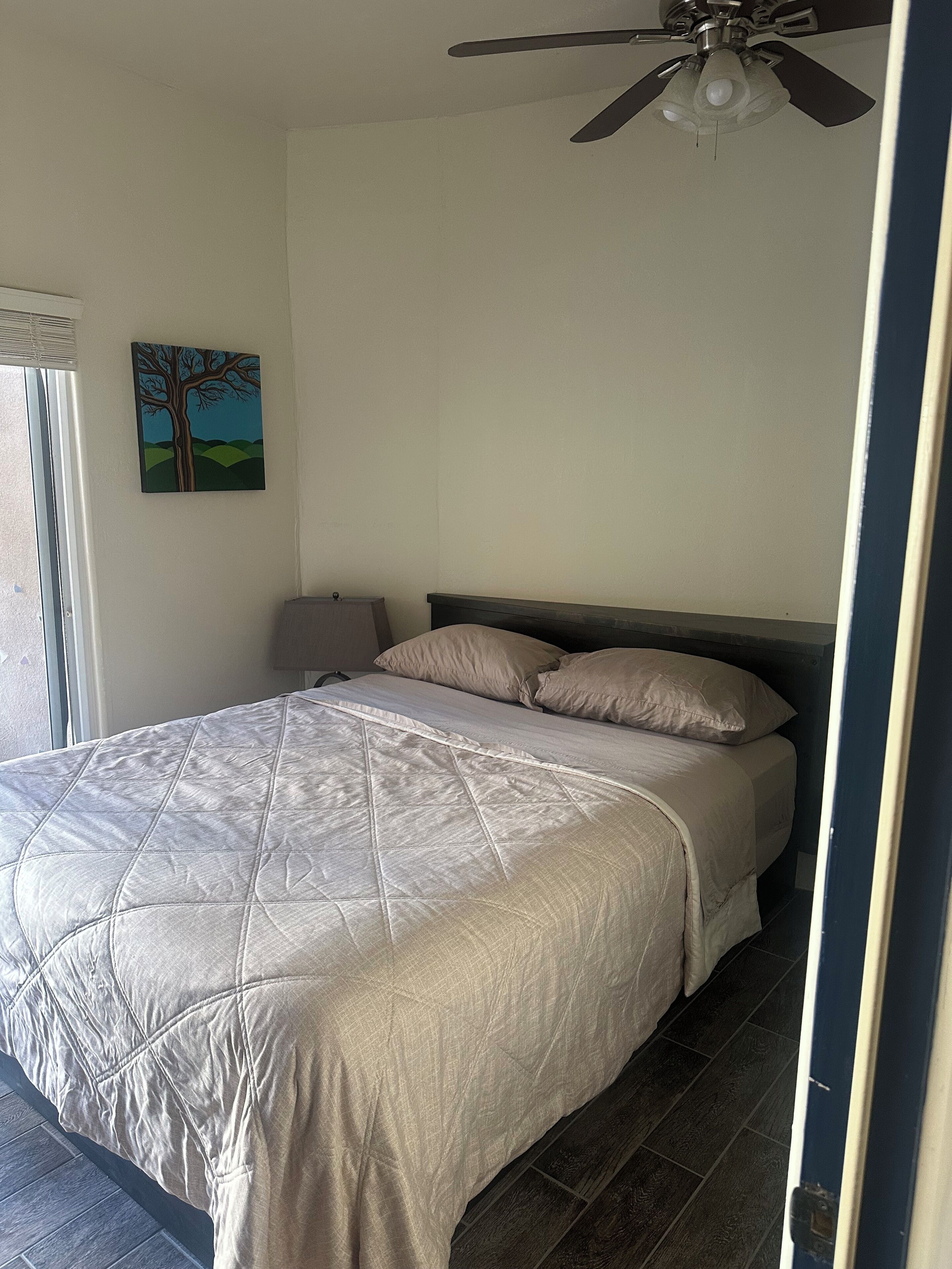 A bedroom with a bed, a ceiling fan, a window, and a wall-mounted painting.