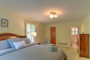 Une chambre avec un grand lit, une table de chevet avec une lampe, une chaise, une armoire en bois et une porte donnant sur une salle de bain.