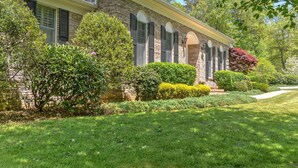 A two-story house with a brick facade, multiple windows with shutters, and a well-maintained front yard with various shrubs and a lawn. - Convenient and spacious 3 br, 1 sofa bed, 2 bath, lg yd, firepit, & patio area. (Asheville)