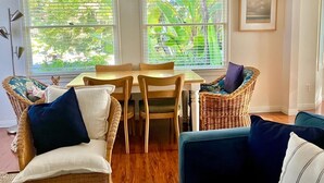 Un comedor con mesa y sillas, ventanas grandes con persianas, muebles de mimbre y un cuadro en la pared.