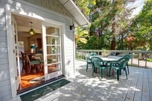 Une terrasse en bois avec une table et des chaises, un patio couvert et une porte donnant sur un espace intérieur agrémenté d’un lustre.