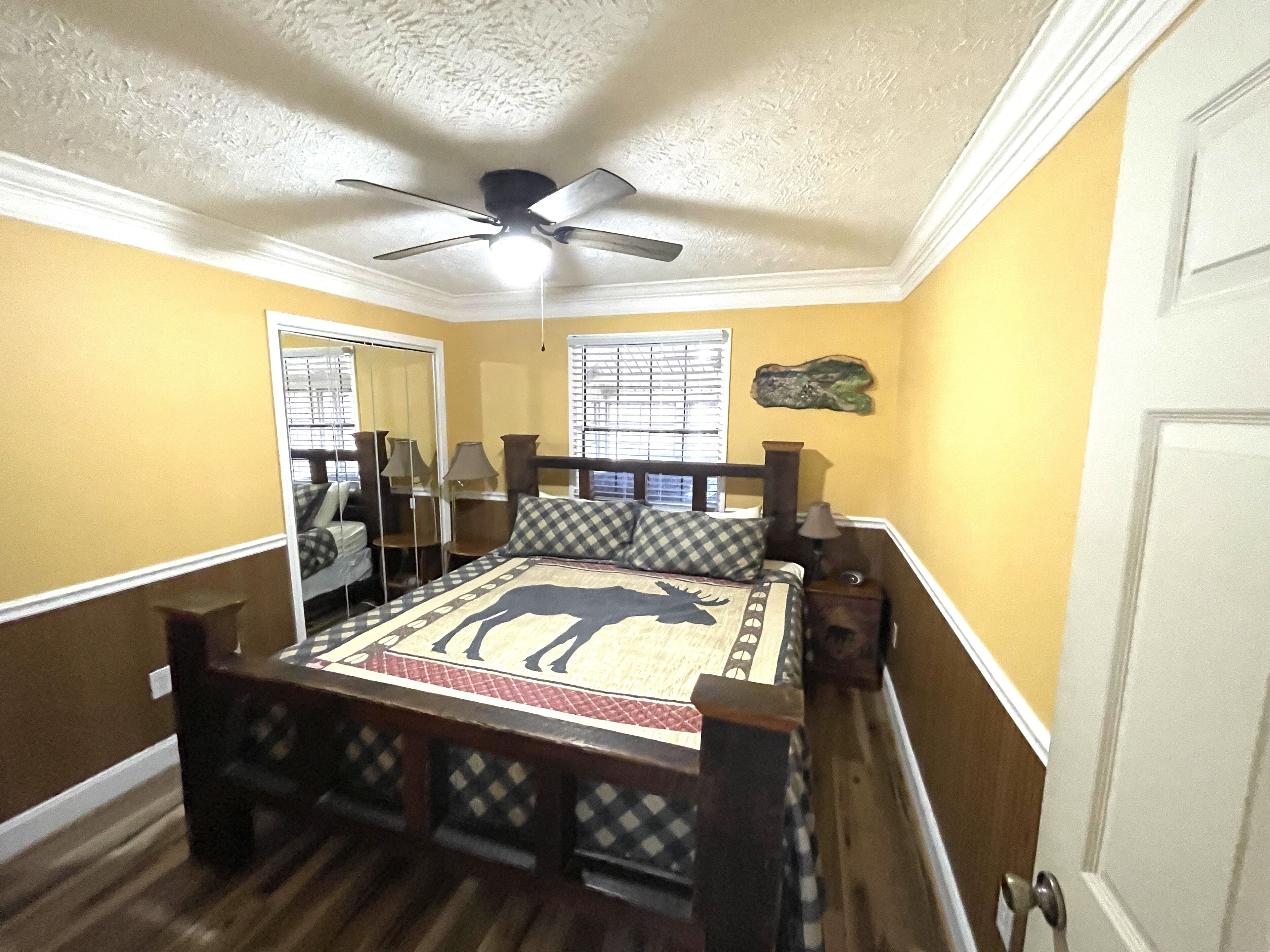 Un dormitorio con cama, ventilador de techo, ventana con persianas, espejo y mesita de noche de madera.