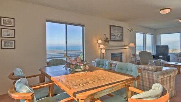 Un salón amplio con una gran mesa de madera, asientos cómodos, chimenea y ventanas grandes con vistas a la playa.