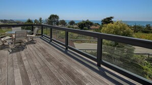 Een houten terras met buitenmeubilair en een glazen balustrade met uitzicht op een woonwijk en de oceaan.