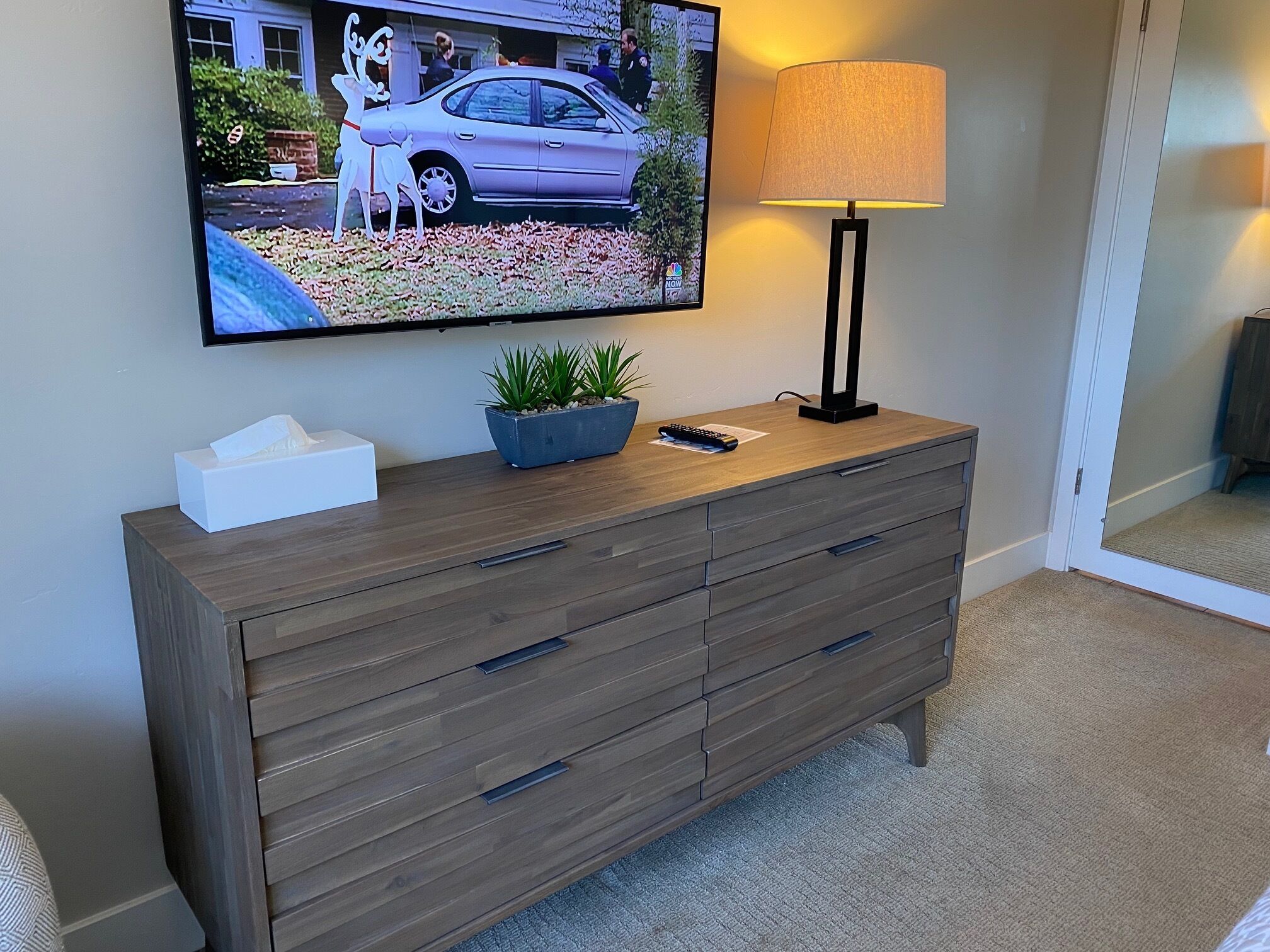 A wooden dresser with drawers, a television mounted on the wall, a lamp, and a small plant on the dresser.
