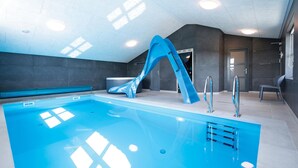 Une piscine intérieure dotée d’un toboggan bleu, d’un jacuzzi et d’un design moderne avec des murs sombres et des luminaires au plafond.