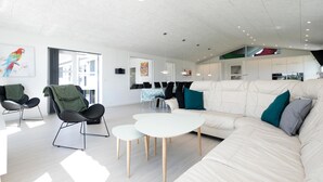 Un vaste espace de vie comprenant un canapé blanc, une table à manger et une chaise noire. Au fond, on aperçoit une cuisine avec des meubles blancs et un appareil électroménager rouge.