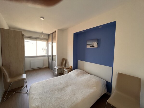 A hotel room with a bed, a desk, a chair, a small table with a lamp, and a window with blinds. - 2-bedroom apartment beachfront with parking, 3 stars (Les Sables-d'Olonne)