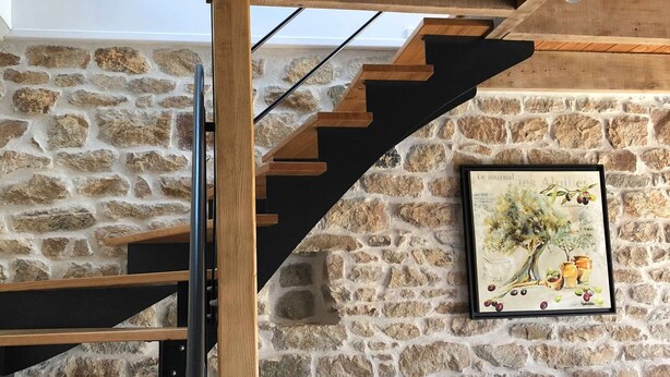 A modern interior with a stone wall, a wooden staircase, a black sofa with mustard cushions, and a framed picture of a tree. - Ty Bihan Small stone house in a quiet location 5 minutes from the beach (FOUESNANT)