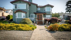 Ein zweigeschossiges Haus mit Veranda, einer roten Tür und einem kleinen Garten mit gelben Blumen und Sträuchern.