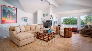 Ein geräumiges Wohnzimmer mit einer großen Eckcouch, einem Glastisch und einem Kamin. Große Fenster sorgen für natürliches Licht und einen Blick ins Freie.
