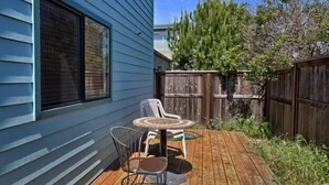 A wooden deck with a round table and two chairs, a wooden fence, and a house with blue siding. - Sea'Scape : 2  BR, 2  BA House in Cambria, Sleeps 4 (Cambria)
