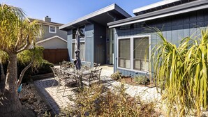 A modern house with a patio area featuring a table and chairs, surrounded by plants and a tree.