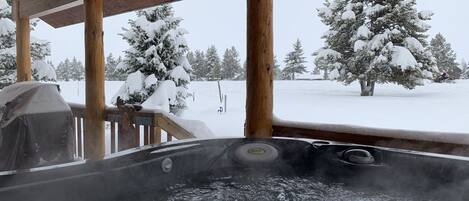 A hot tub under a wooden deck with a snowy landscape view.