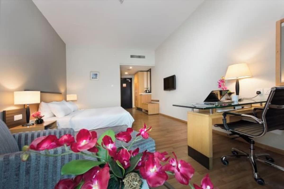 Habitación de hotel con una cama, un escritorio con computadora, una silla, una lámpara y un florero con flores.
