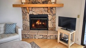 Ett vardagsrum med en öppen spis i sten, en tv på ett träställ och en soffa med ett mönstrat kuddöverdrag.