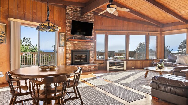 A spacious living area with a fireplace, a television mounted on a brick wall, and large windows offering a view of the outdoors. - A Cool House, A Cool View (Cambria)