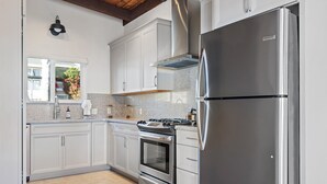 A modern kitchen with white cabinets, stainless steel appliances, and a window with a view of a garden. - A Cool House, A Cool View (Cambria)