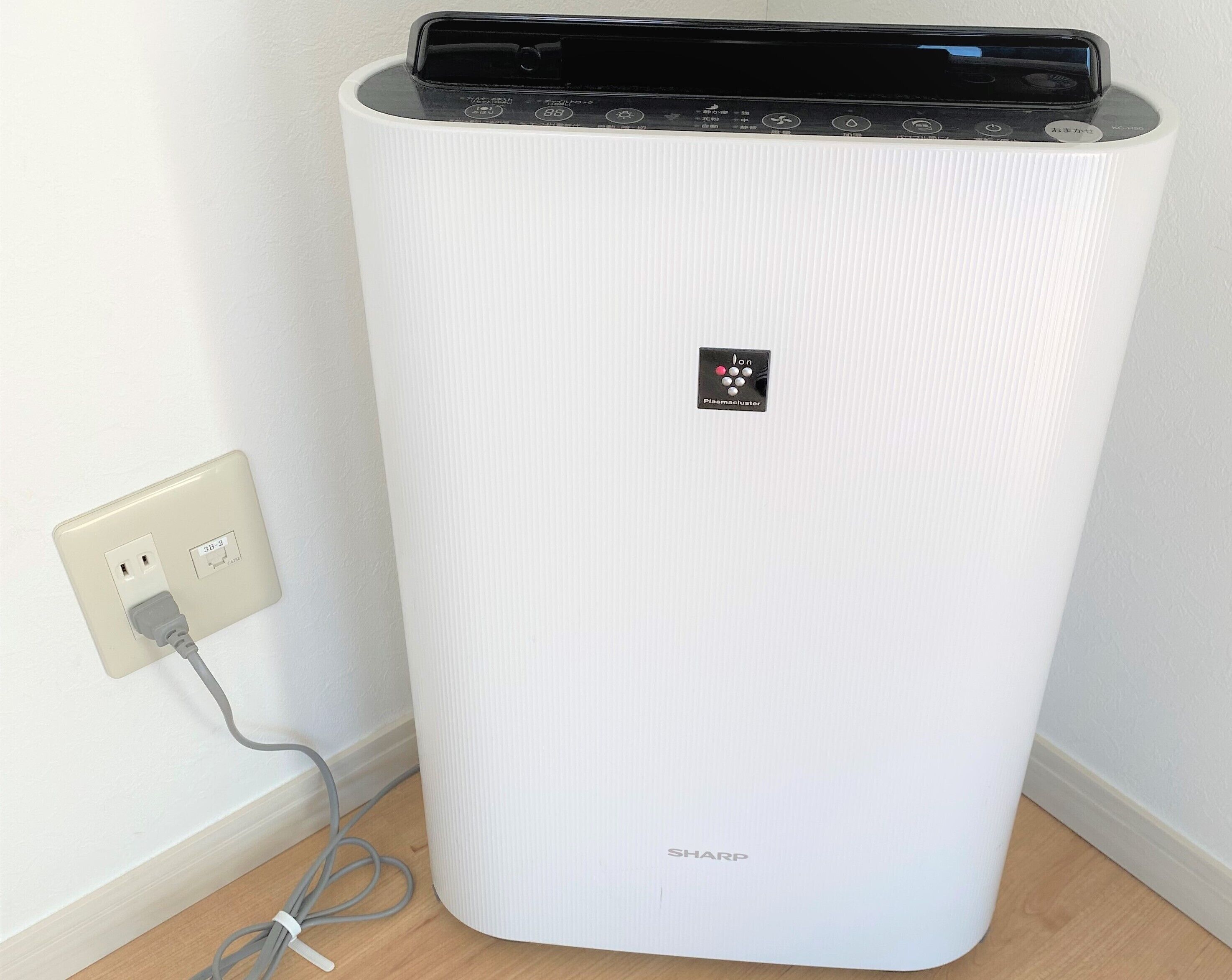 A white, ribbed, rectangular air purifier with a control panel and a power cord plugged into a wall socket.