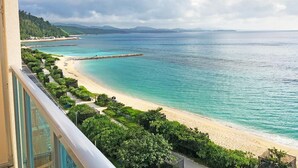 Vista costeira de uma varanda com vista para uma praia, um paredão rochoso, vegetação exuberante e montanhas ao longe.