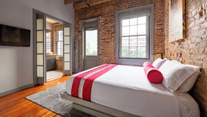 Een slaapkamer met een bakstenen muur, een bed met witte beddengoed en een rode kussen, een raam en een schilderij aan de muur.