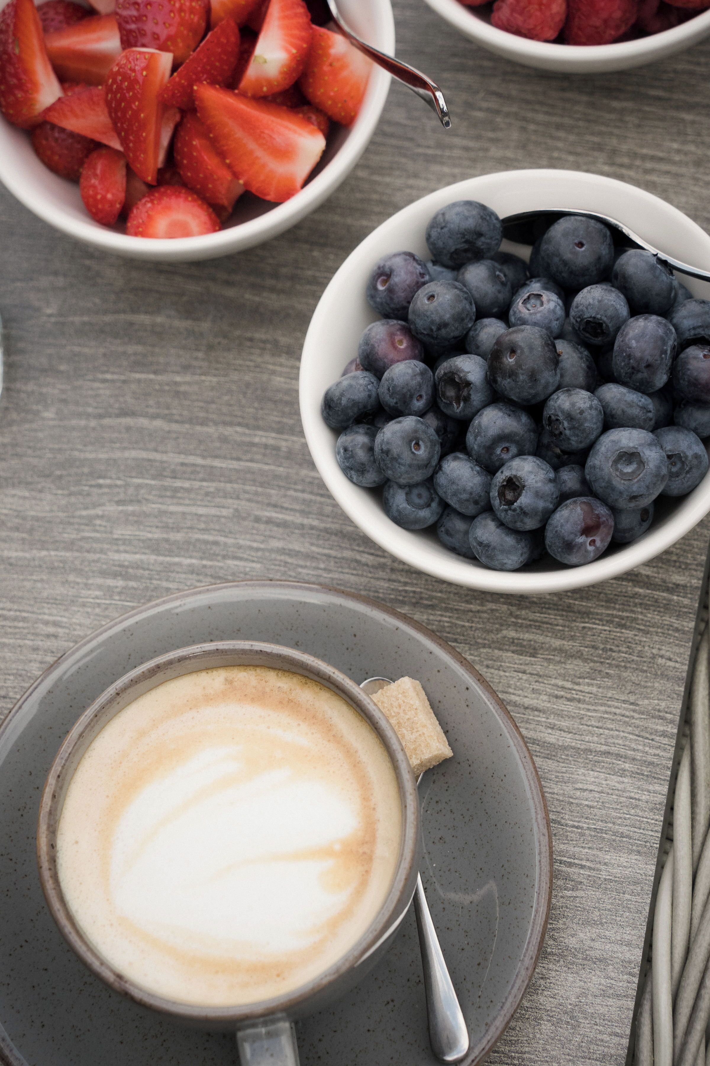 A table with a cup of coffee, a bowl of strawberries, and a bowl of blueberries.