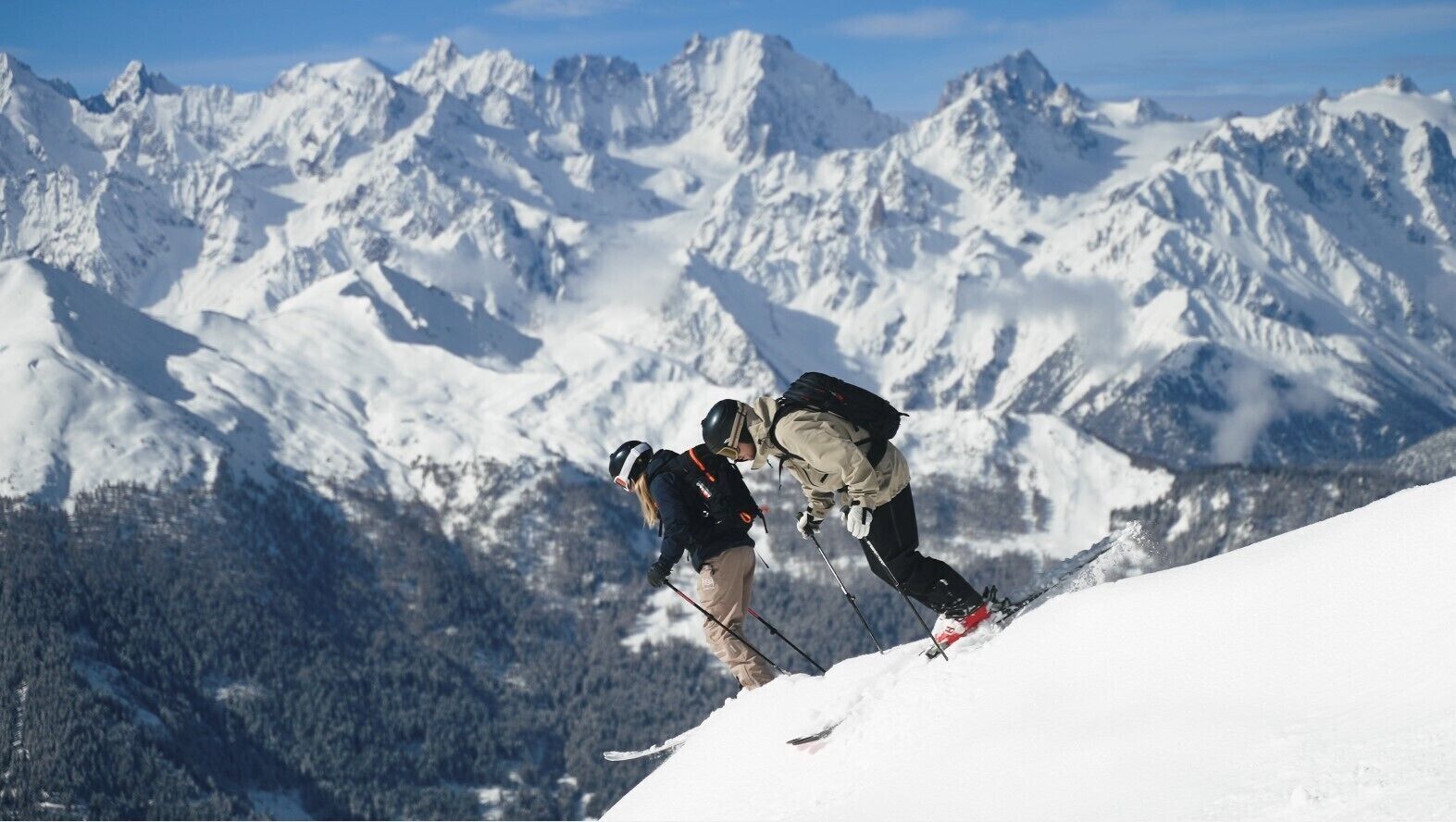Two skiers on a snowy mountain slope with a backdrop of snow-covered peaks.