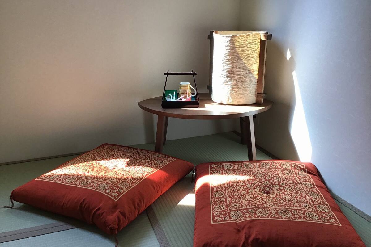 Una habitación con esteras de tatami, dos cojines rojos con motivos florales, una mesita con una lámpara y una pila de libros.
