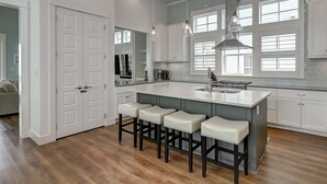 Una cocina moderna con una isla grande, taburetes de bar, iluminación colgante y vista al área de estar.