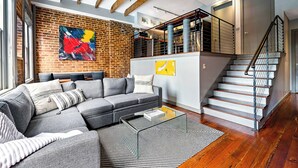 A modern living room with a sectional sofa, a coffee table, and a large abstract painting on the brick wall. - Private Balconies w/ Heated Pool, Hot Tub Near French Qtr – Family Friendly (New Orleans)