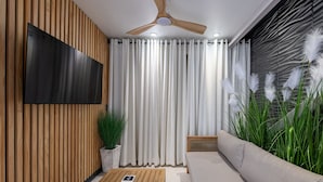 Un salon moderne doté d’un téléviseur à écran plat, de murs recouverts de panneaux de bois, d’un ventilateur de plafond et d’une grande fenêtre ornée de rideaux légers.