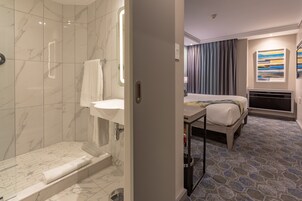 A modern bathroom with a glass-enclosed shower, a sink, and a toilet. The room features a bed with a wooden frame, a bedside table, and a patterned floor.