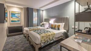 Habitación de hotel con dos camas, un escritorio, una silla, un televisor y un ventanal grande con cortinas.