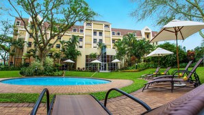 A hotel pool area with lounge chairs and umbrellas, surrounded by greenery and a building with multiple windows. - StayEasy Mbombela (Mbombela)