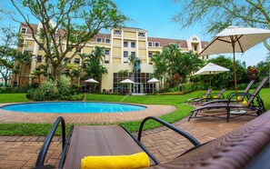 A hotel pool area with lounge chairs and umbrellas, surrounded by greenery and a building with multiple windows. - StayEasy Mbombela (Mbombela)