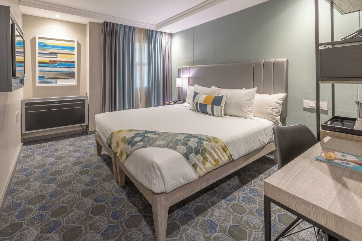 A hotel room with a large bed, a desk, a chair, a television, and a painting on the wall.
