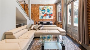 A modern living room with a sectional sofa, a glass coffee table, and a large abstract painting on the brick wall. - Poolside 3BR w Hot Tubs, Grill Near French Qtr, Great for Families & Groups (New Orleans)